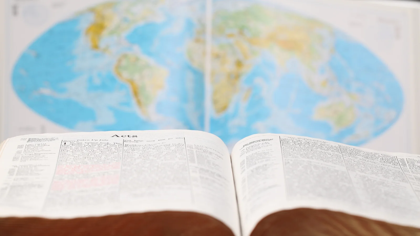 uma Bíblia aberta no livro de Atos com um mapa do mundo ao fundo